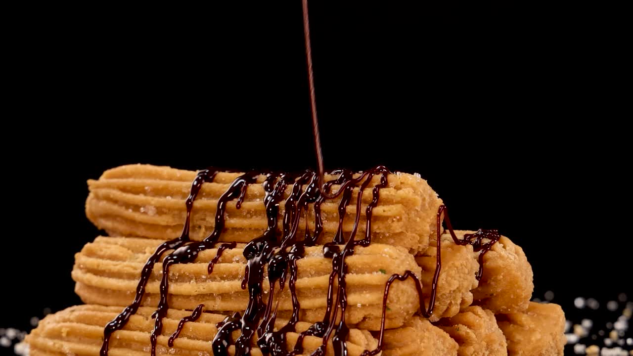 Golden churros are stacked on a dark background as rich chocolate sauce is poured over them in slow motion, highlighting texture and indulgence