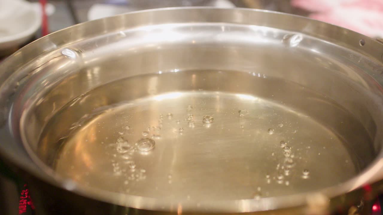 Hand places morning glory greens into simmering hotpot broth under bright, even kitchen lighting