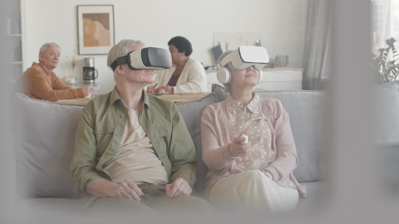 Medium long of grey-haired man and woman wearing virtual reality headsets, sitting on couch in living room, moving heads sideways, up and down, talking, smiling