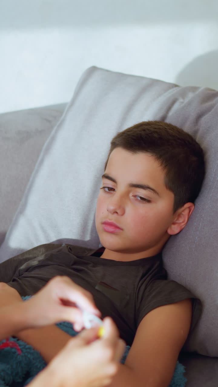 Girl with ponytail and maroon shirt checks thermometer reading after removing it from sick boy resting on couch showing care concern and responsibility in calm indoor space with soft light
