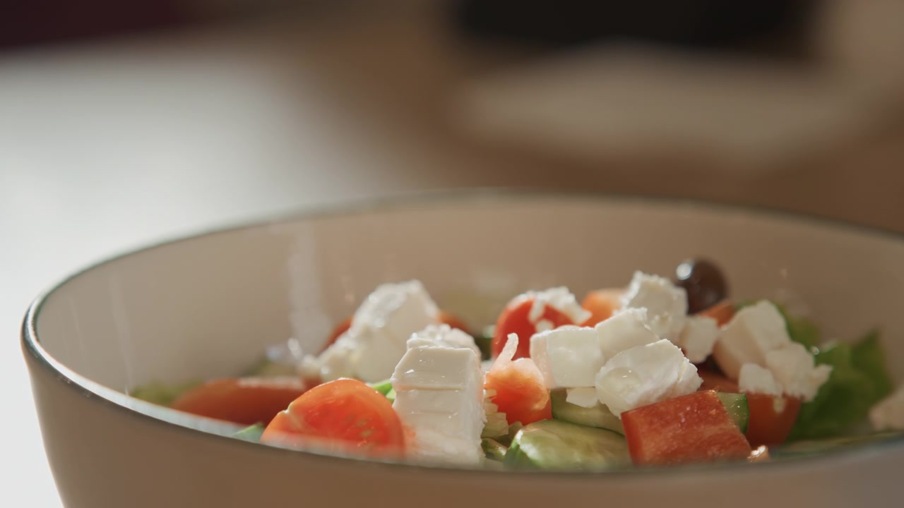 Primo piano di ingredienti freschi per insalata, inclusi pomodori, cetrioli, formaggio feta e olive in una ciotola. L'illuminazione soffusa esalta i colori vivaci su uno sfondo interno con tonalità calde.