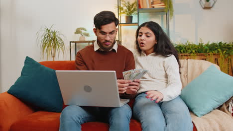 happy indian couple counting money and booking tickets for holiday vacation sitting on sofa at home