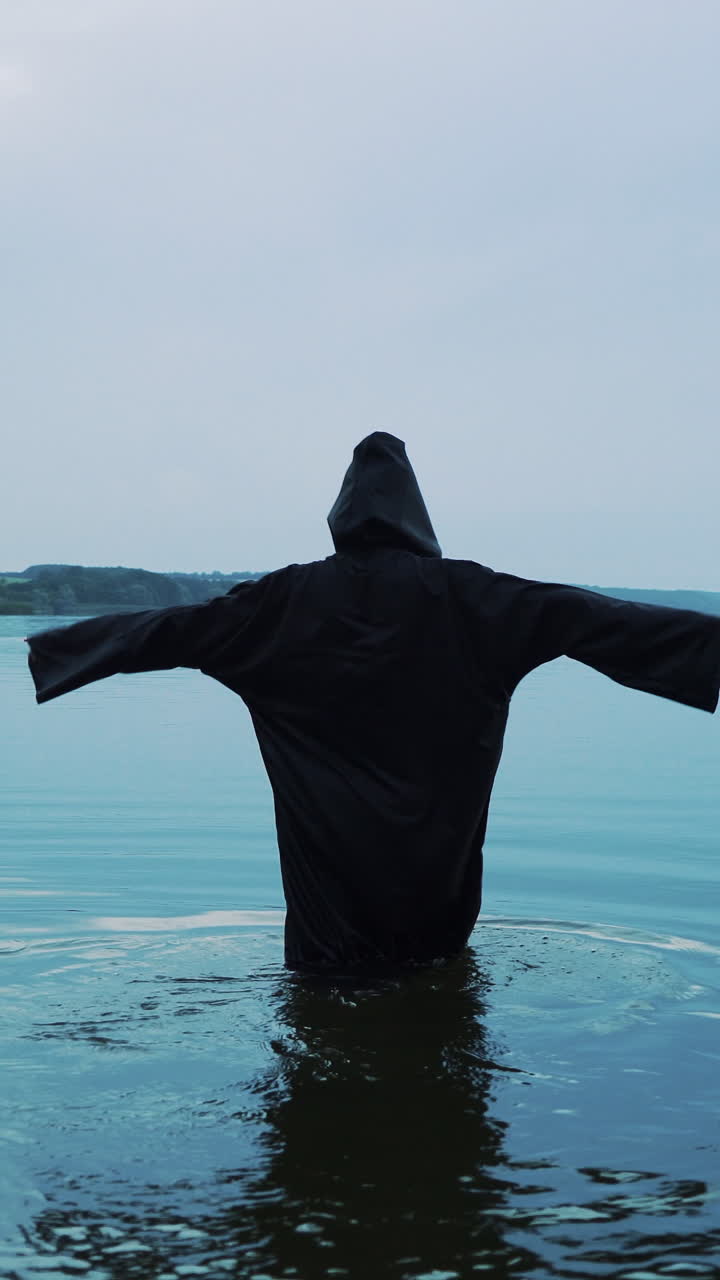 Female figure in a black robe enters the water. Halloween costume. Vertical video