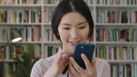 close up portrait of beautiful cute asian woman standing in library using smartphone smiling laughing bookcase in background