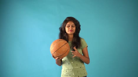Ein-Junges-Indisches-Mädchen-In-Grünem-T-Shirt,-Das-Mit-Basketball-In-Der-Hand-Spielt-Und-In-Die-Kamera-Schaut,-Die-In-Einem-Isolierten-Studio-Mit-Blauem-Hintergrund-Steht