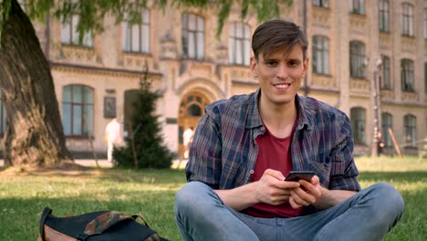 joven apuesto está sentado en la hierba en el parque, tocando en el teléfono inteligente, wayching en la cámara, concepto de relajación, concepto de comunicación, la construcción en el fondo