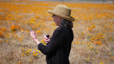 Eine-Alte-Frau,-Die-Auf-Einem-Smartphone-SMS-Schreibt-Und-Versucht,-Mobilfunkempfang-Zu-Bekommen,-Während-Sie-In-Zeitlupe-Auf-Einem-Naturspaziergang-In-Einem-Blumenfeld-Unterwegs-Ist