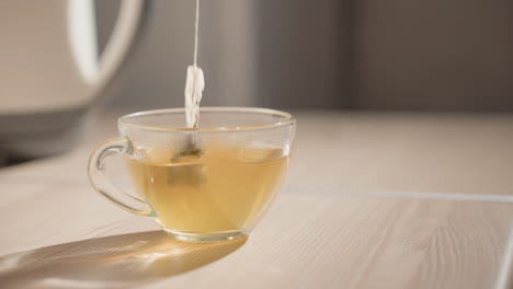 Close-up of clear glass cup with tea bag steeping. Light golden tea in cup with steam rising. Soft lighting highlights the serene and cozy atmosphere in minimalist kitchen
