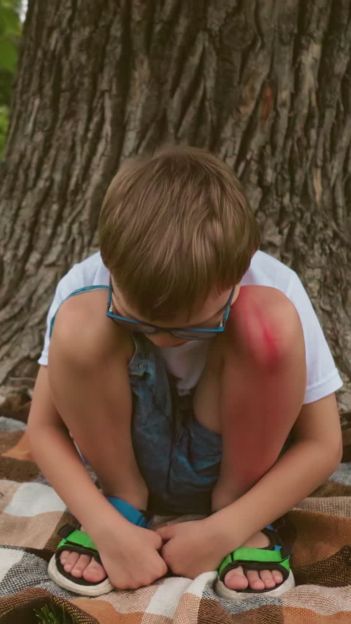 uma criança pequena senta-se debaixo de uma grande árvore, profundamente pensativa com uma contusão visível na perna, as mãos juntas, e uma bicicleta azul está deitada no chão perto