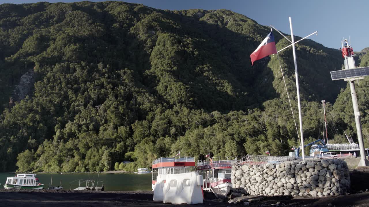 Chilean flag waving on a lakeside pier in Petrohué, surrounded by lush green mountains and boats on calm water. Peaceful scene symbolizing adventure and national identity