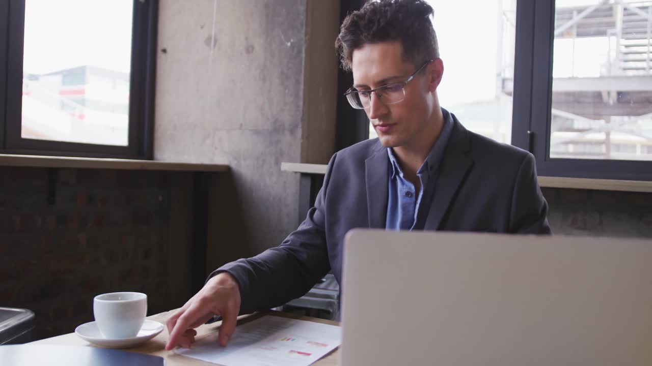 Uomo d'affari caucasico che usa un computer portatile e esamina documenti, seduto a un tavolo con un caffè in un bar.