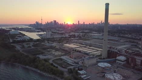 aerial sunset wide shot flying over industrial factory past huge smokestack tower with downtown skyline in background in toronto ontario canada