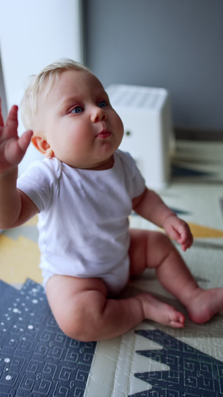 Adorable plump Caucasian baby wearing white bodysuit sits on the floor near the window. Little kid puts his hands to his mouth and looks up. Vertical video.