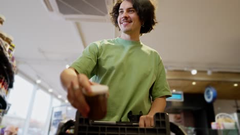 Bottom-view-of-a-happy-brunette-guy-with-curly-hair-in-a-green-T-shirt-puts-necessary-goods-in-a-cart-during-his-shopping-in-a-supermarket