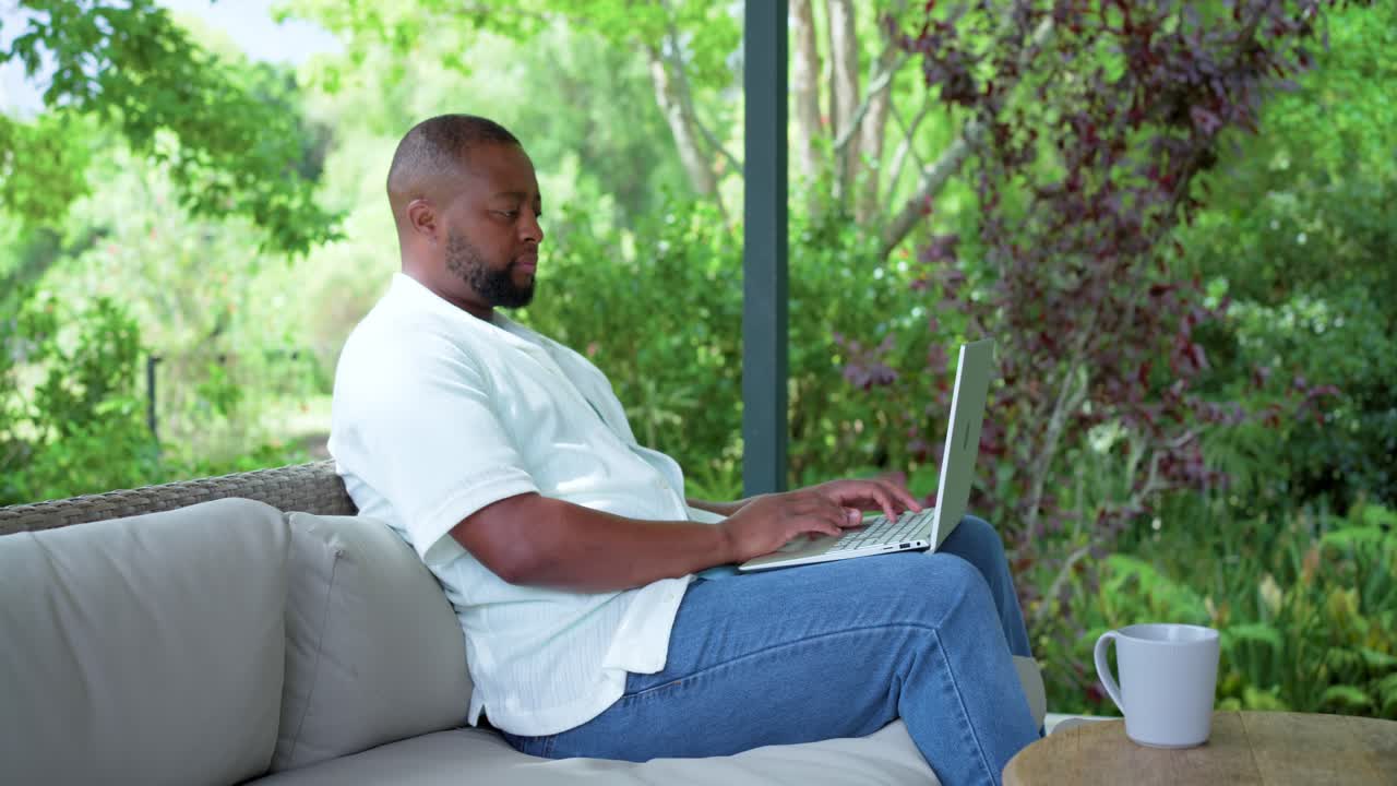 Adult African American man on porch sofa, typing on laptop while responding to work, mug nearby