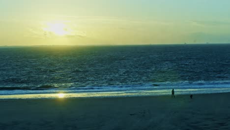 Blick-Auf-Das-Meer-Und-Den-Strand-Am-Abend-Bei-Sonnenuntergang,-Während-Ein-Mann-Mit-Einem-Hund-Im-Abendrot-Mit-Wellen,-Wasser-Und-Sonne-Im-Weitwinkelpanorama-Spazieren-Geht