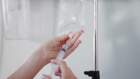 Hands-of-caucasian-female-doctor-filling-drip-bag-from-syringe-in-hospital-ward,-slow-motion
