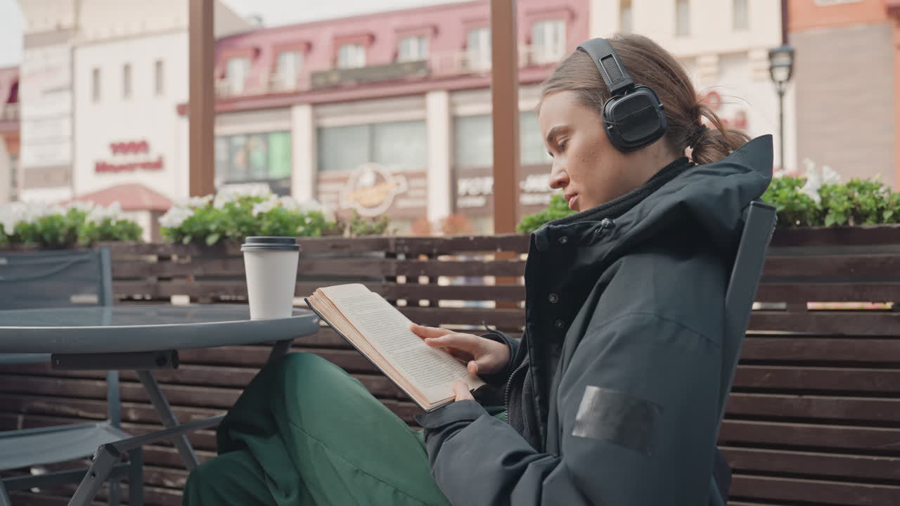 Темнокожая женщина читает книгу в уличном кафе в наушниках. На столе — теплая куртка и чашка кофе. На заднем плане — витрина городского магазина. Расслабленное, задумчивое выражение лица, медленное перелистывание страниц, утренний свет, уютная неспешная жизнь