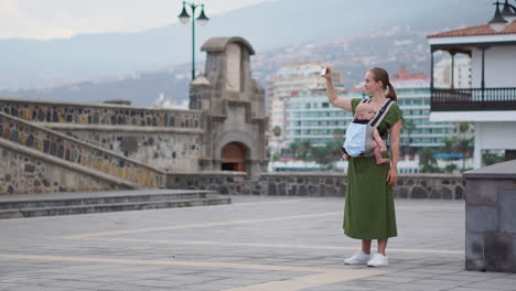 a young woman on a journey has her baby in a kangaroo backpack and uses a mobile phone to take pictures. as they walk, she checks the screen periodically