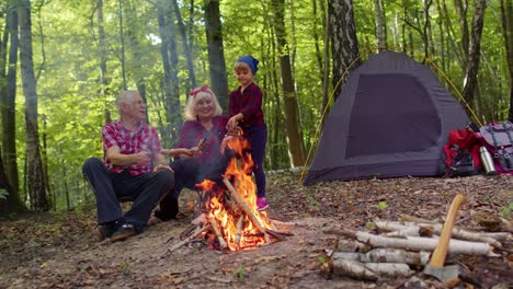 Senior-grandmother-grandfather-with-granddaughter-cooking-frying-sausages-over-campfire-in-wood