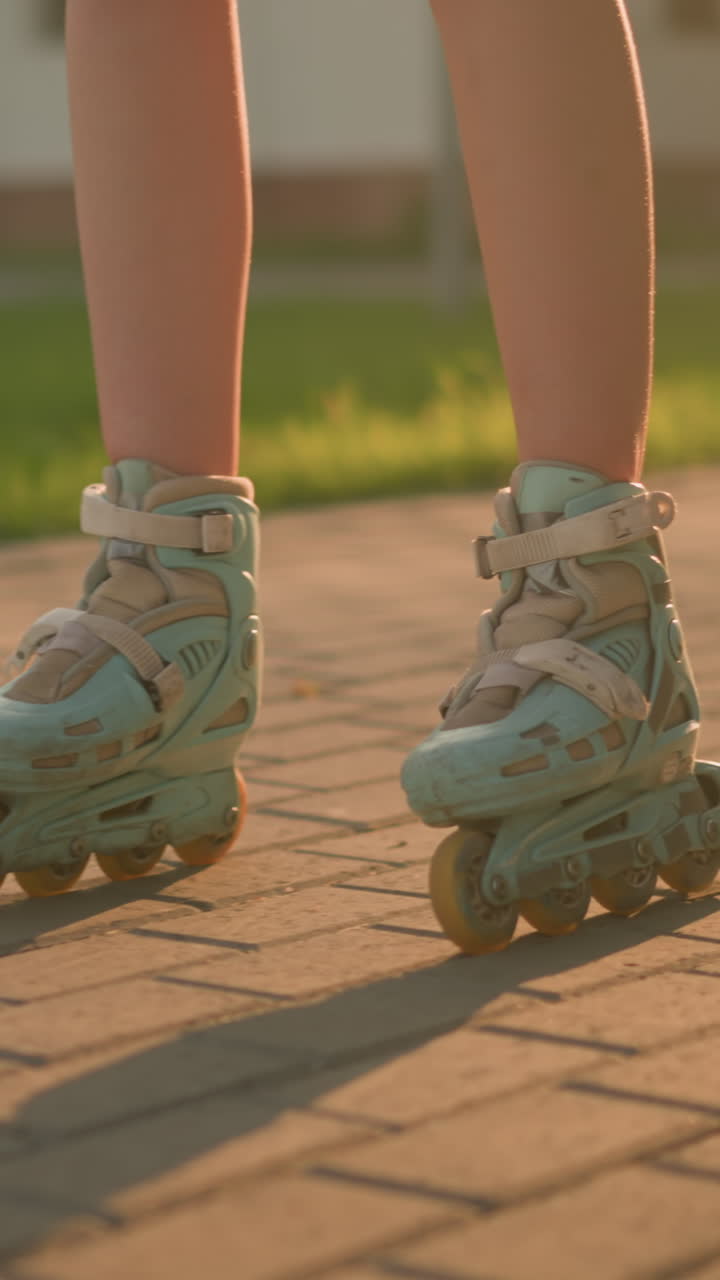 Leg view of lady roller skating slowly on interlocked path, wearing teal rollerblades, casting shadow on ground under warm sunlight, blurred background featuring lush greenery, building and trees