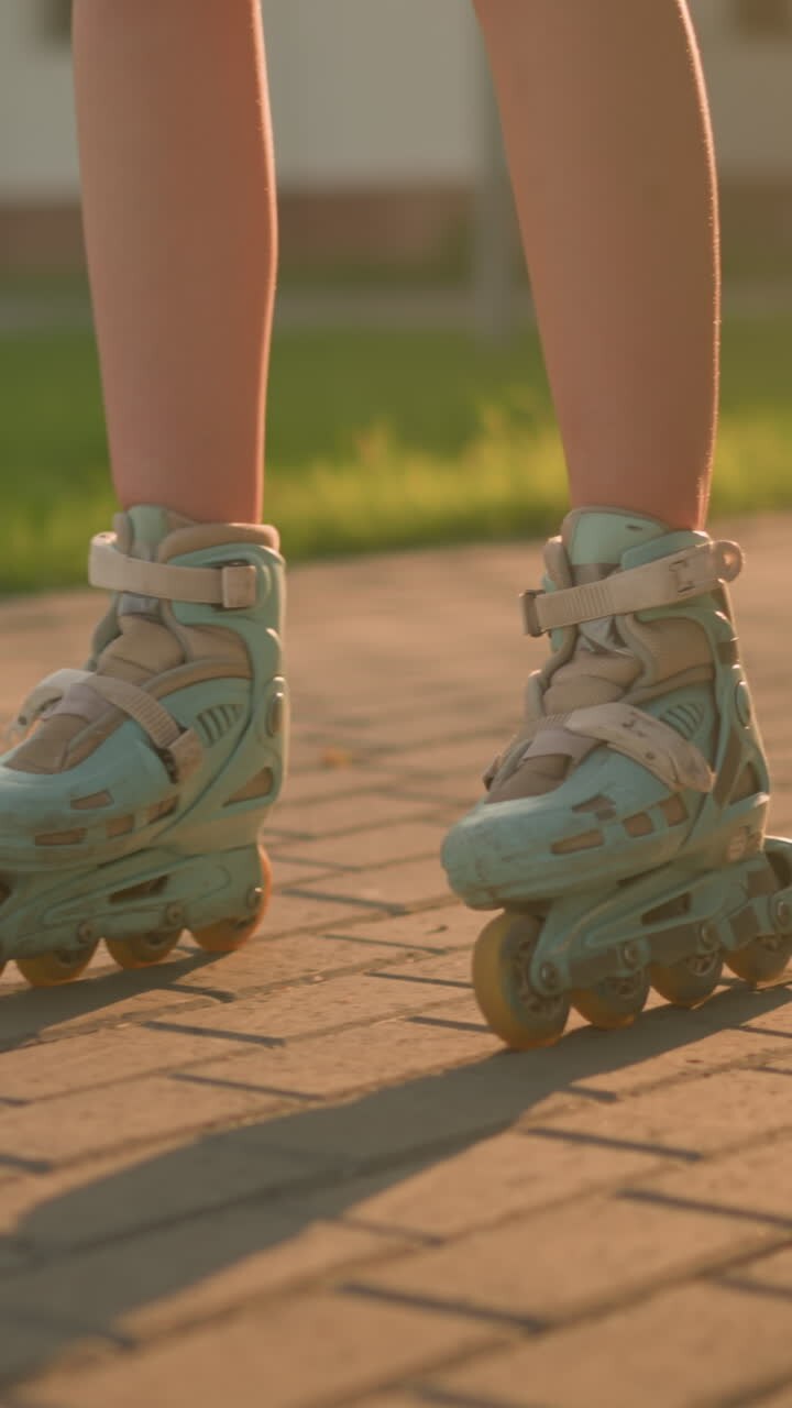 Leg view of lady roller skating slowly on interlocked path, wearing teal rollerblades, casting shadow on ground under warm sunlight, blurred background featuring lush greenery, building and trees