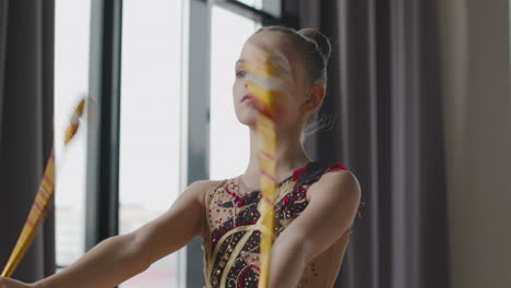 focused young girl in leotard practising rhythmic gymnastics with clubs in a studio