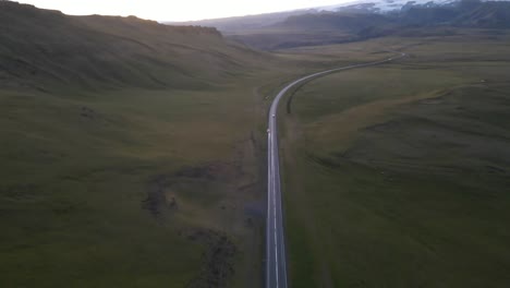 Capture-La-Tranquilidad-Del-Atardecer-Con-Un-Dron-Deslizándose-Sobre-Una-Carretera-Solitaria-Mientras-Un-Automóvil-Atraviesa-La-Vasta-Vegetación-Sin-árboles-De-Islandia,-Abrazado-Por-Siluetas-De-Montañas-En-La-Suave-Luz.