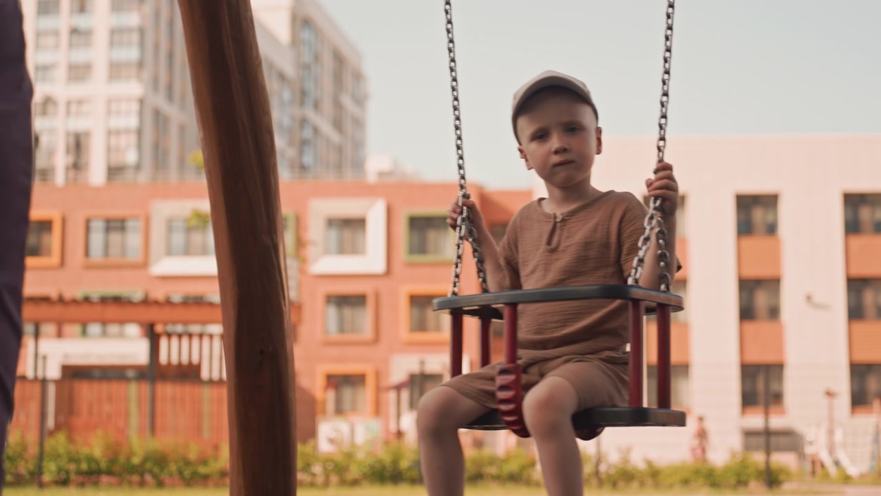Un giovane ragazzo si diverte a dondolarsi al parco giochi, indossando abiti casual e tenendosi alle catene. Una donna adulta sta nelle vicinanze in un ambiente di parco urbano con edifici visibili sullo sfondo sotto la luce del giorno.