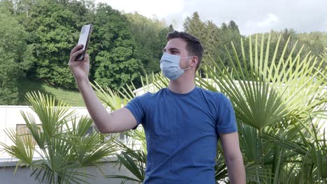 young man in blue shirt makes selfies in the garden while wearing his face mask