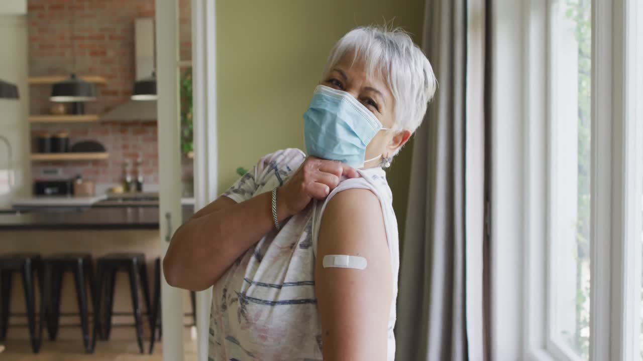 Happy mixed race senior woman in face mask showing plaster on arm after covid vaccination