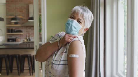 happy mixed race senior woman in face mask showing plaster on arm after covid vaccination