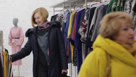 two elderly women go into a fashion boutique and choose clothes hanging on hangers.