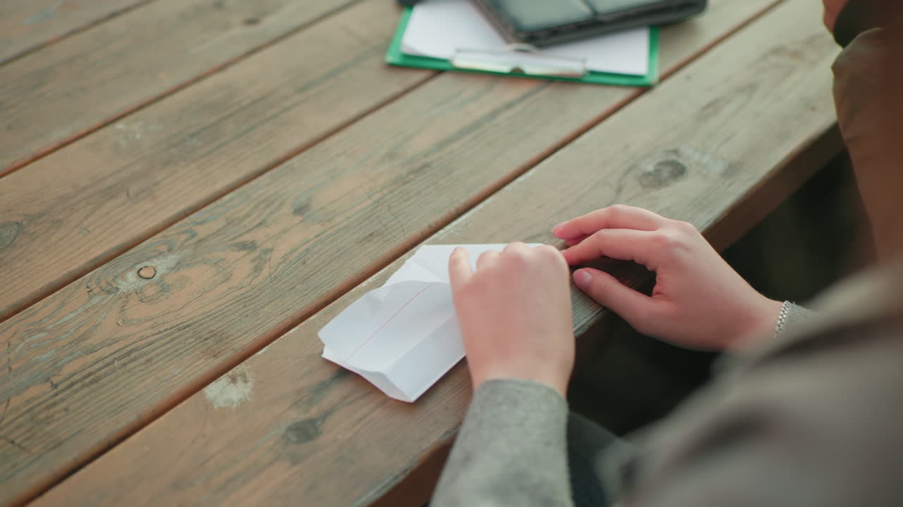 Close up view of person folding paper into kite on wooden table with notebook in background, showing detailed hand movement during crafting process in casual outdoor setting