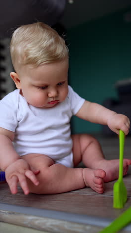 fair-haired infant baby in white bodysuit sits on the floor. little kid plays with kitchen brush. vertical video.
