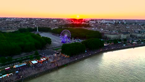 Ferris-Wheel-in-Place-des-Quinconces-Bordeaux-France,-rotating-and-illuminated-in-Neon-during-sunset,-Aerial-pan-right-shot