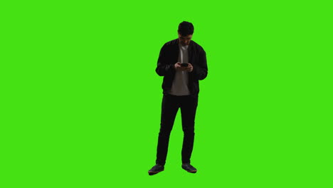 joven enviando o leyendo un mensaje de texto o desplazándose en línea en un teléfono móvil de pie contra una pantalla verde con iluminación de clave baja 1