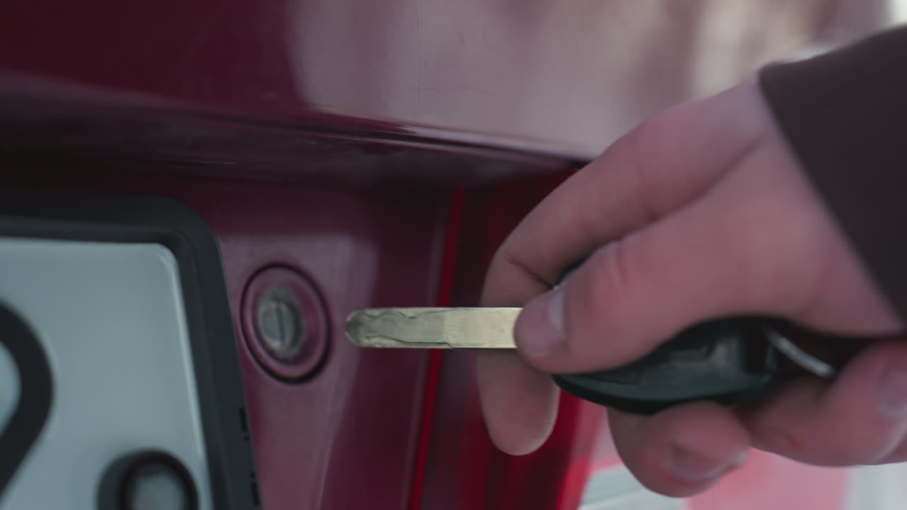 close up of car bonnet latch area as person approaches key inserted into lock to open hood, glossy paint reflections under cold outdoor light with blurred background hinting at winter scene