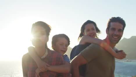 Portrait-Of-Parents-Giving-Children-Piggybacks-By-The-Sea