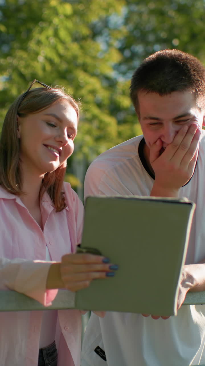 dama de camiseta rosa mostrando a su hermano algo de su tableta mientras él se ríe cubriendo su boca, ambos apoyados en la barandilla en el parque al aire libre con luz solar suave y vegetación exuberante