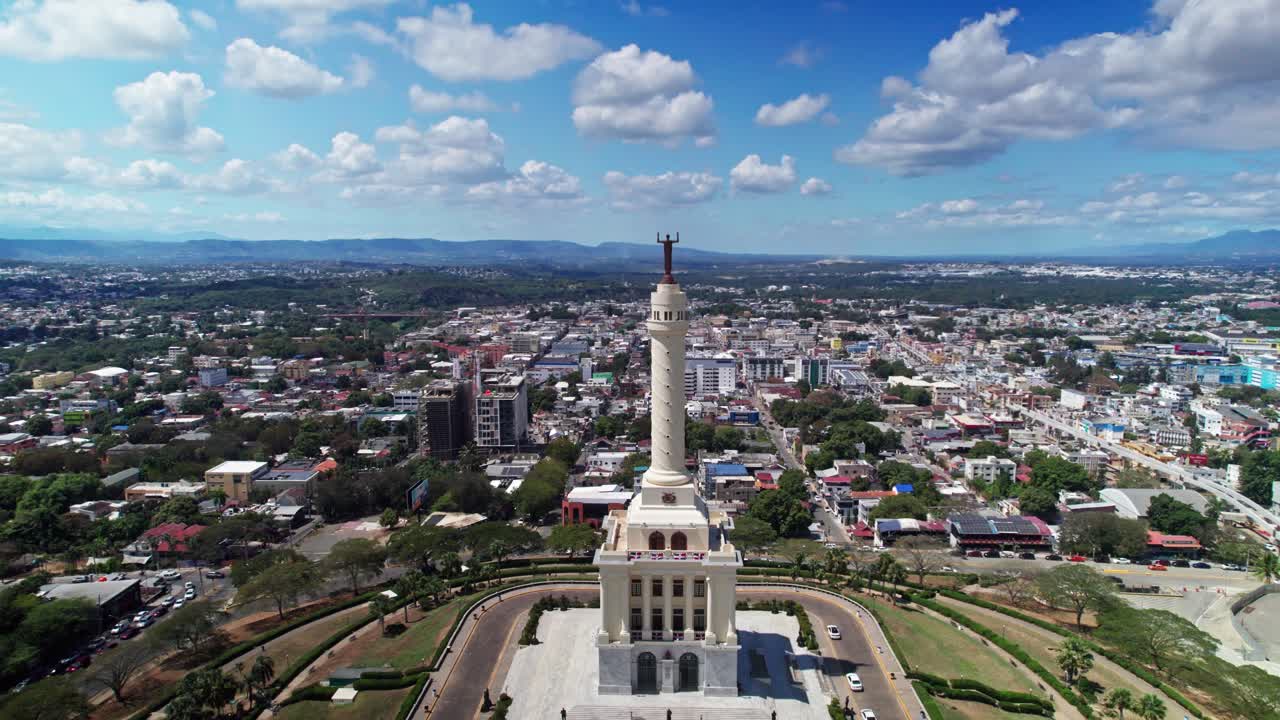 Ripresa aerea con drone in allontanamento che rivela il Monumento agli Eroi della Restaurazione. Ampia vista aerea del punto di riferimento architettonico e dello skyline della città di Santiago sotto un cielo azzurro brillante.