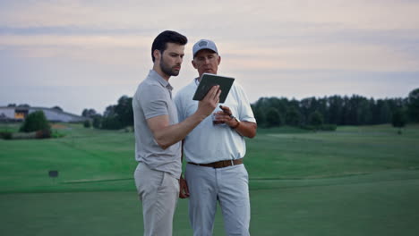 two golfers looking tablet watching tournament game video at golf course sunset.