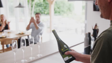 Ein-älterer-Mann-öffnet-Eine-Flasche-Champagner,-Während-Die-Familie-Mit-Erwachsenen-Nachkommen-Zu-Hause-Am-Tisch-Isst