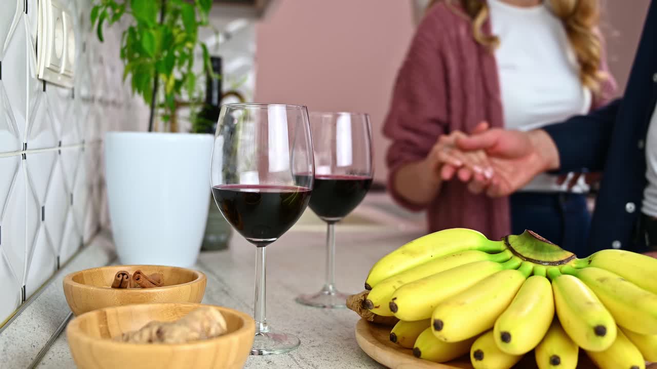 Man's hand caress an woman hands in a white modern kitchen on blurred background. Glasses with red wine, bananas, cinnamon and ginger on wooden tables in foreground. Valentines day in love