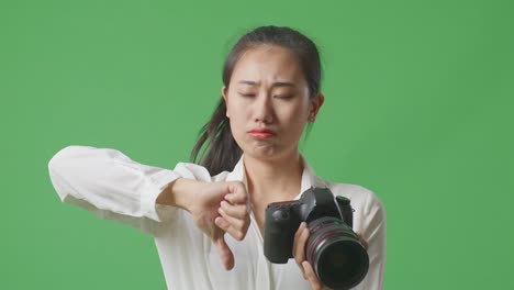 close up of upset asian photographer looking at the pictures in the camera then showing thumbs down gesture while standing on green screen background in the studio