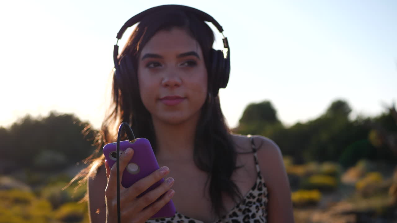 una hermosa joven hispana sosteniendo un teléfono inteligente escuchando música feliz en los auriculares al aire libre bajo la épica luz del sol con bengalas en cámara lenta