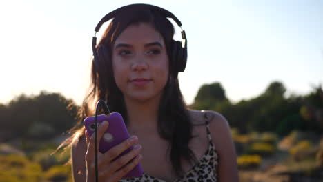 a beautiful young hispanic woman holding a smartphone listening to happy music on headphones outdoors in epic sunlight with lens flares in slow motion