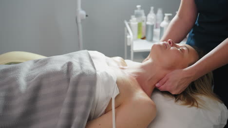 close up of professional gliding fingertips along side of client neck toward shoulder during relaxing spa treatment on soft linens in bright clinic room with organized product trolley in background