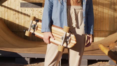 video of midsection of caucasian female skateboarder holding skateboard in skate park