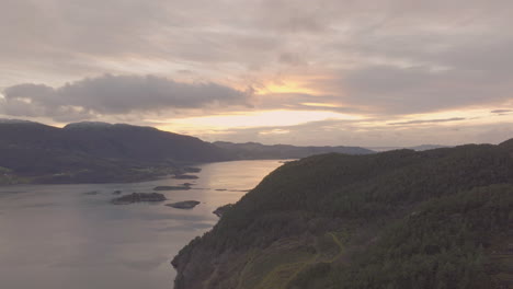 voo aéreo panorâmico sobre a montanha arborizada ao lado do fiorde, pôr do sol de fogo